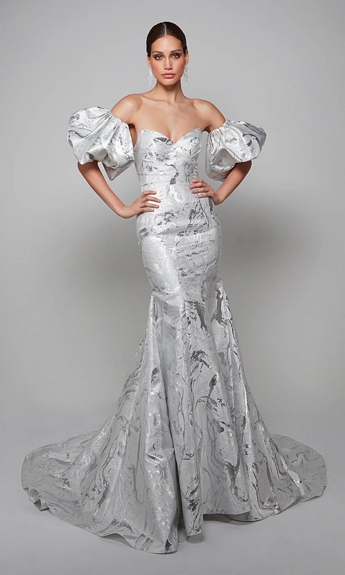 A woman poses in the Long White Dress: Alyce 7055, an off-the-shoulder mermaid gown with dramatic puff sleeves, ideal for a modern mother-of-the-bride look, set against a plain gray background.
